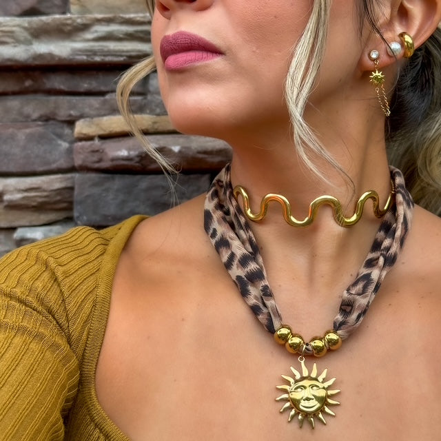 Woman wearing a gold necklace with a sun pendant and a leopard print scarf, against a stone wall background.