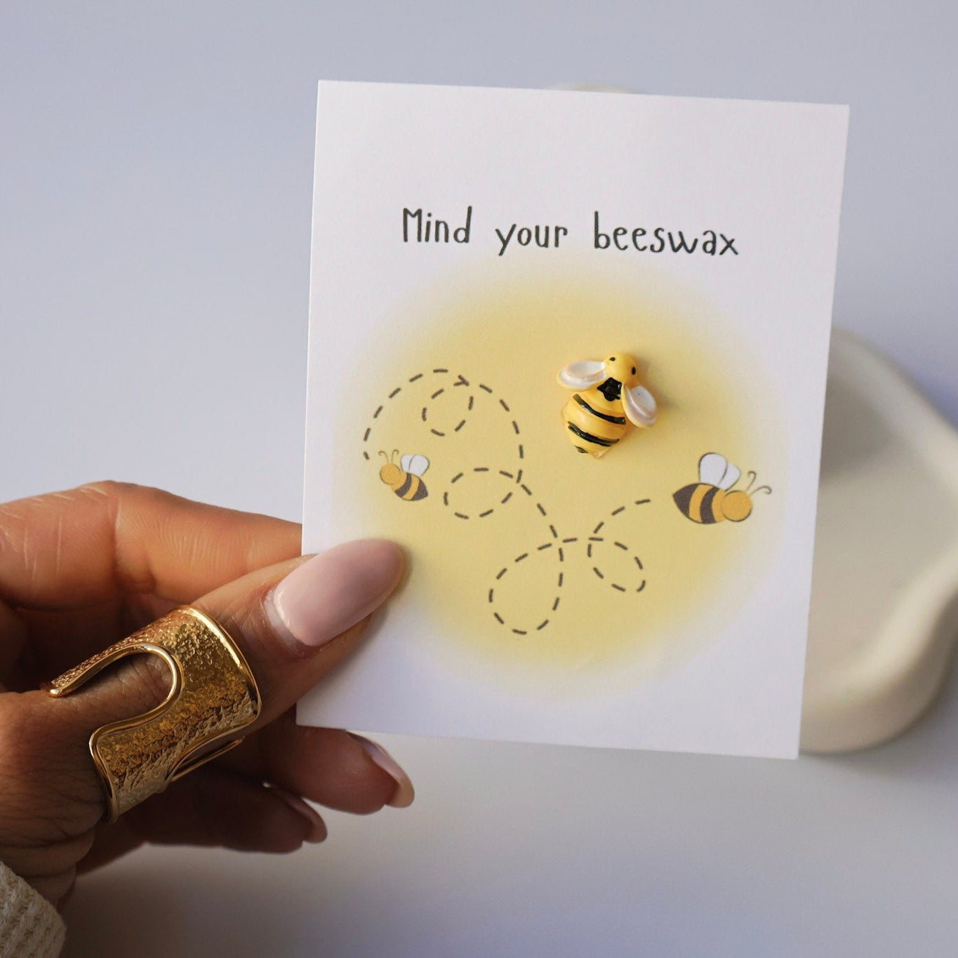 Hand holding a card with bee illustrations and 'Mind your beeswax' text on a light background