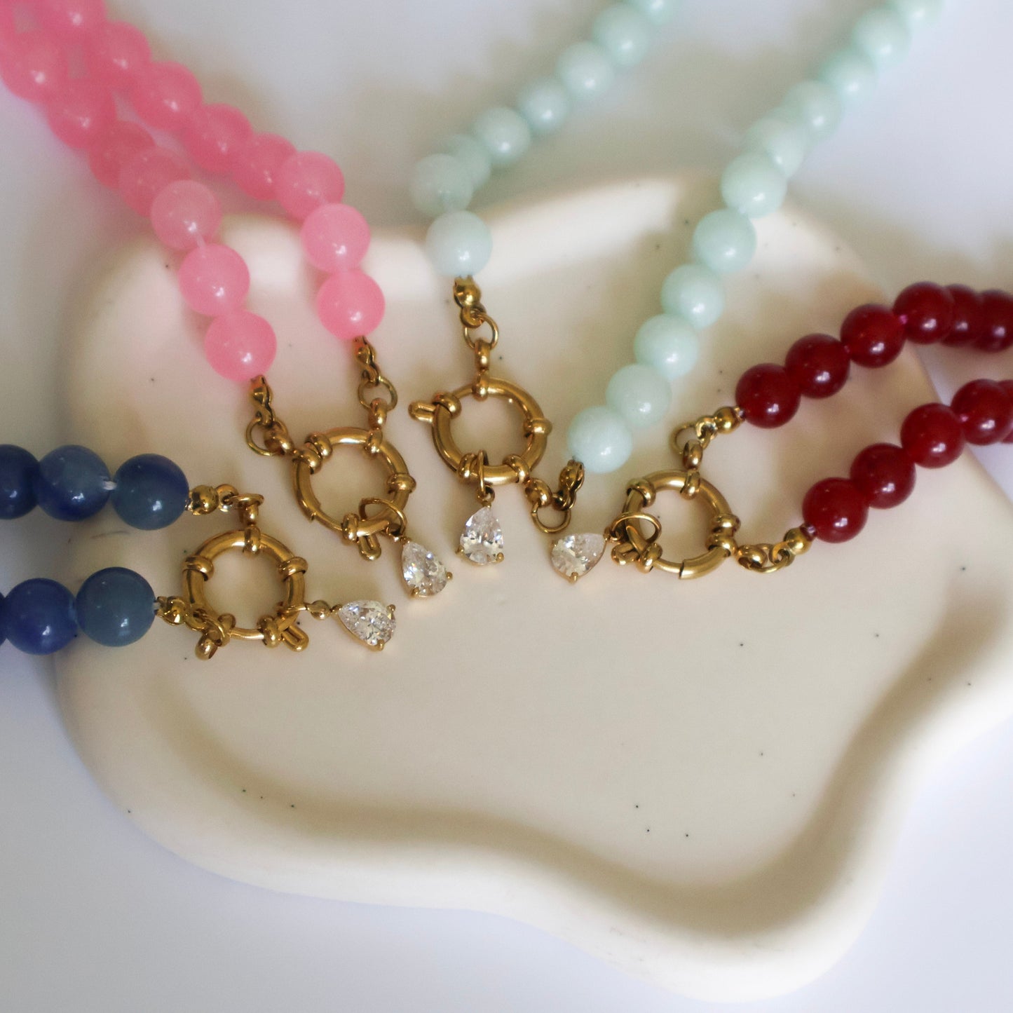 Four beaded necklaces in pink, blue, green, and red on a white surface.