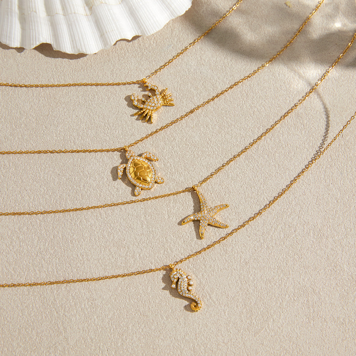 Four delicate gold chain necklaces with sea-themed pendants, including a crab, turtle, starfish, and seahorse, arranged diagonally on a textured beige surface with a white seashell in the corner.
