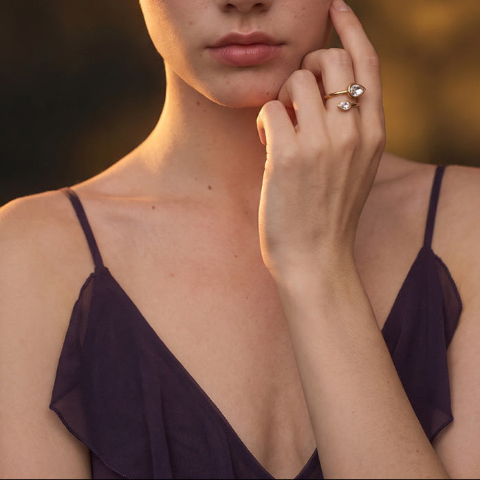 Model wearing the Radiance Double Crystal Ring, an adjustable 18K gold-plated ring featuring two clear teardrop-shaped crystals, styled with a purple dress in warm sunlight.