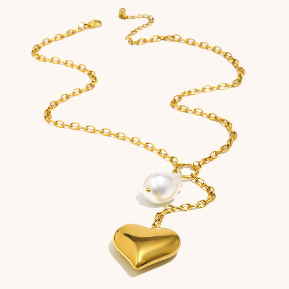 A gold chain necklace featuring a polished gold heart pendant and a small white pearl bead, displayed on a clean off-white background.