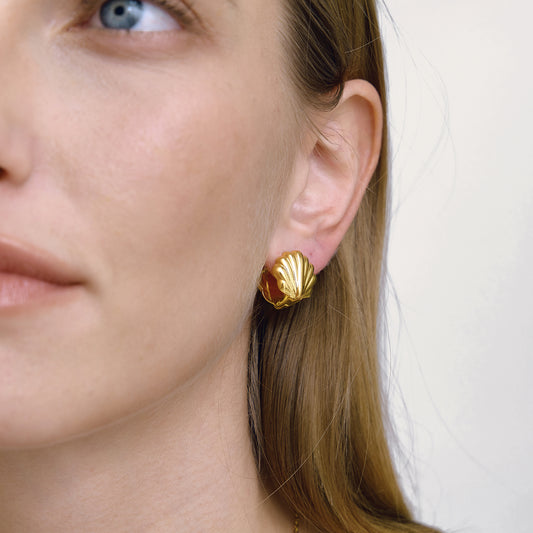 Close-up of a person wearing gold shell-shaped earrings against a neutral background