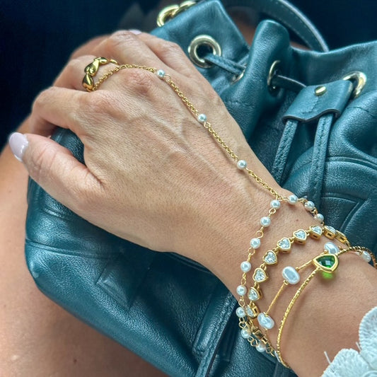 Hand wearing gold bracelets with pearls and a green gemstone, holding a teal handbag.