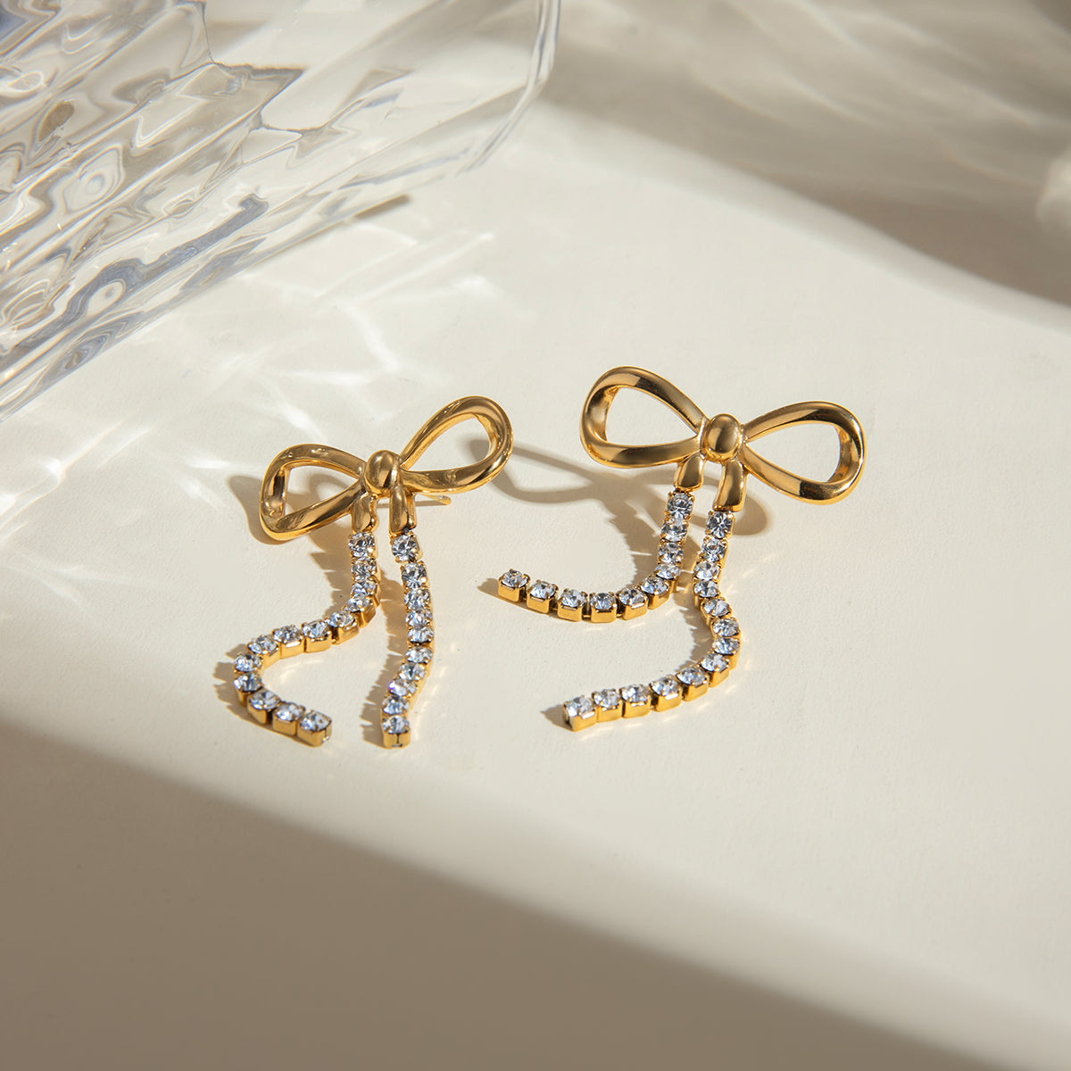 Gold bow-shaped earrings with clear gemstones on a beige surface