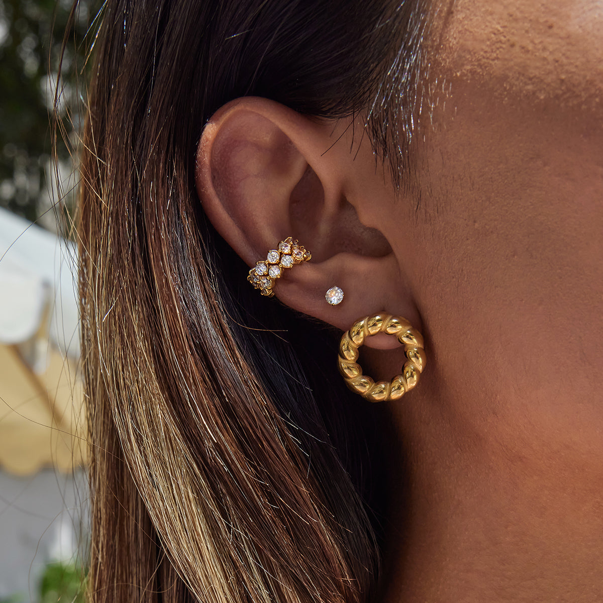 Close-up of an ear wearing gold hoop earrings with a blurred outdoor background