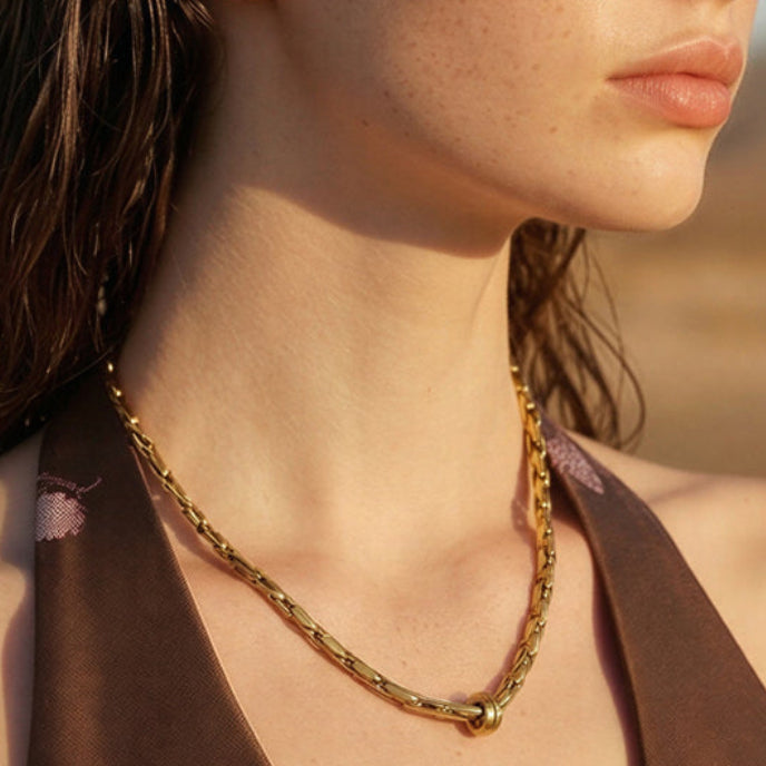The Covenant gold-plated woven chain necklace worn with a brown halter top, showing the polished ring detail below the collarbone