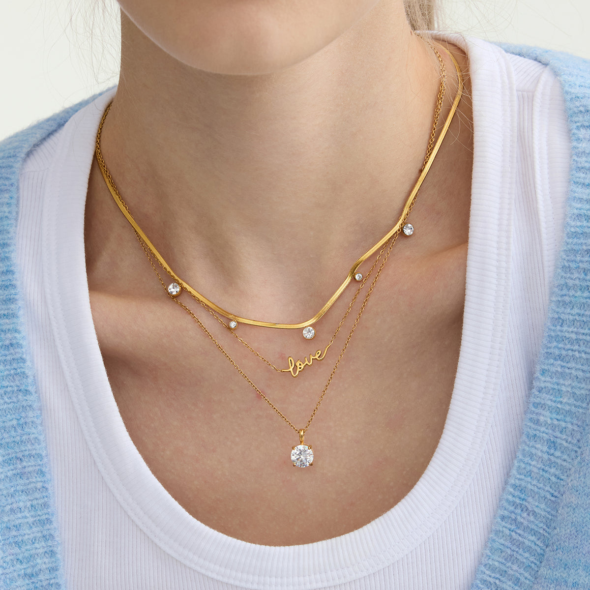 Gold necklaces with small stones and a 'love' pendant on a person wearing a white top.