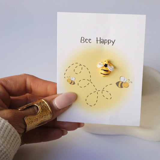 Hand holding a card with bee illustrations and 'Bee Happy' text on a light background