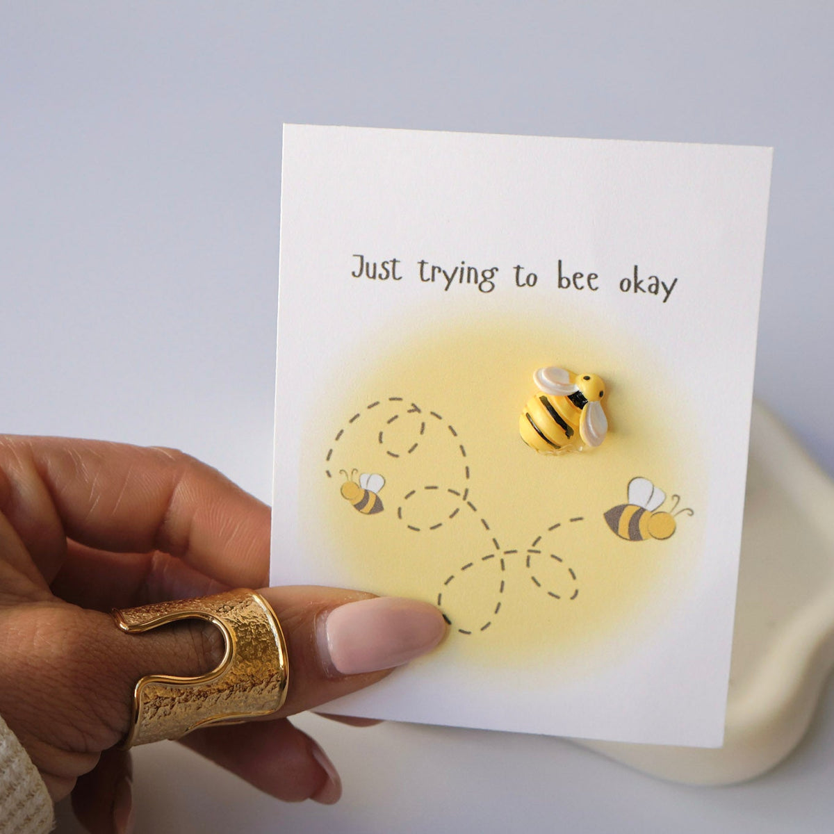 Hand holding a card with bee illustrations and text 'Just trying to bee okay' on a light background.