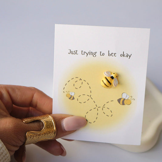 Hand holding a card with bee illustrations and text 'Just trying to bee okay' on a light background.