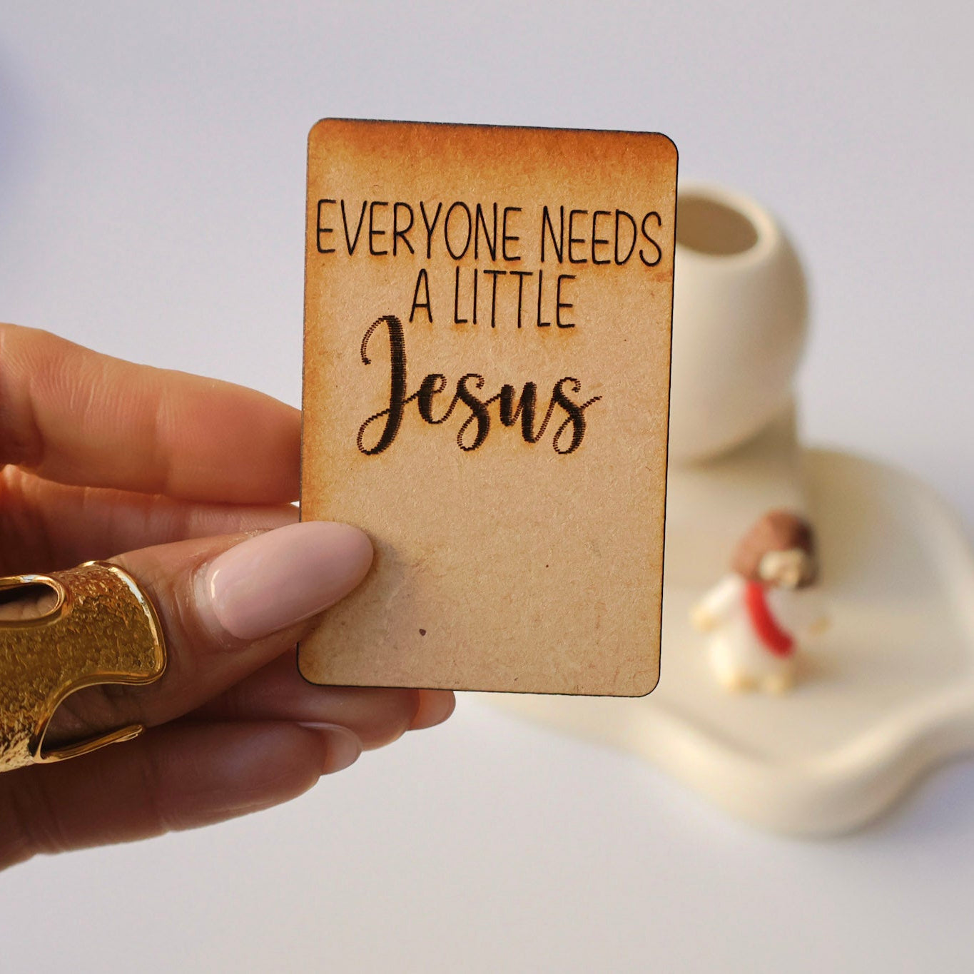 Hand holding a wooden card with 'Everyone needs a little Jesus' text, with a blurred background.