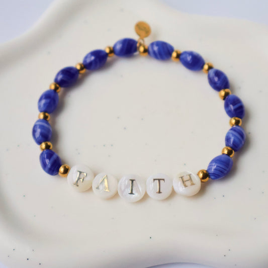 Blue beaded bracelet with letter beads spelling 'FAITH' on a white dish.