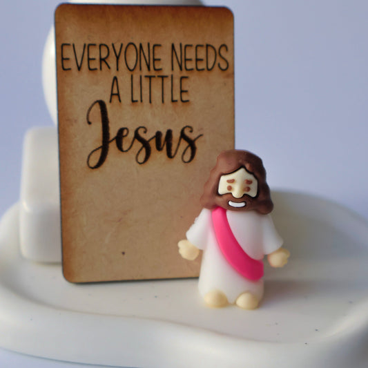 Small figurine of a person with a pink tunic next to a wooden sign with 'Everyone needs a little Jesus' text on a white base.