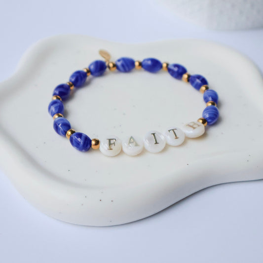 Bracelet with blue beads and letter beads spelling 'FAITH' on a white dish.