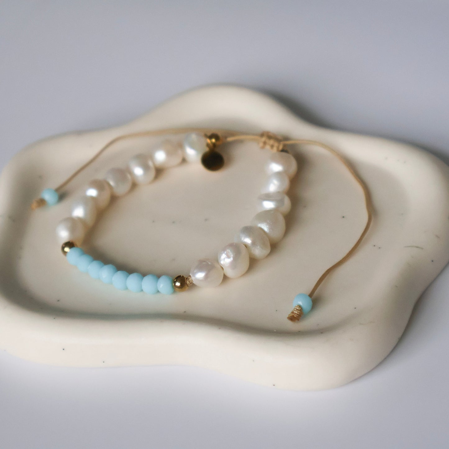 Two pearl bracelets with turquoise and gold accents on a white ceramic dish.