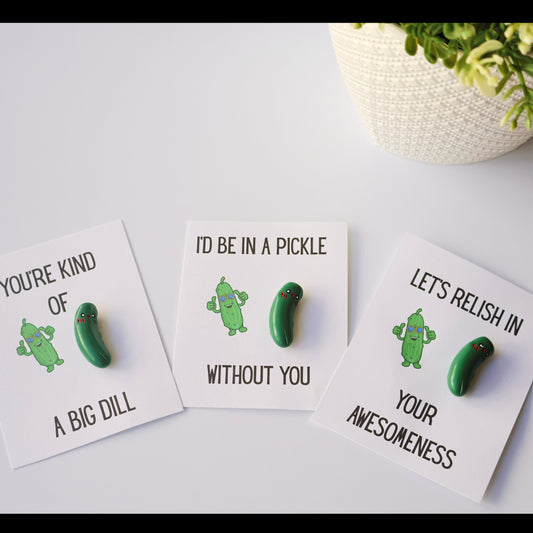 Three cards with pickle-themed charms on a light surface with a plant in the background.