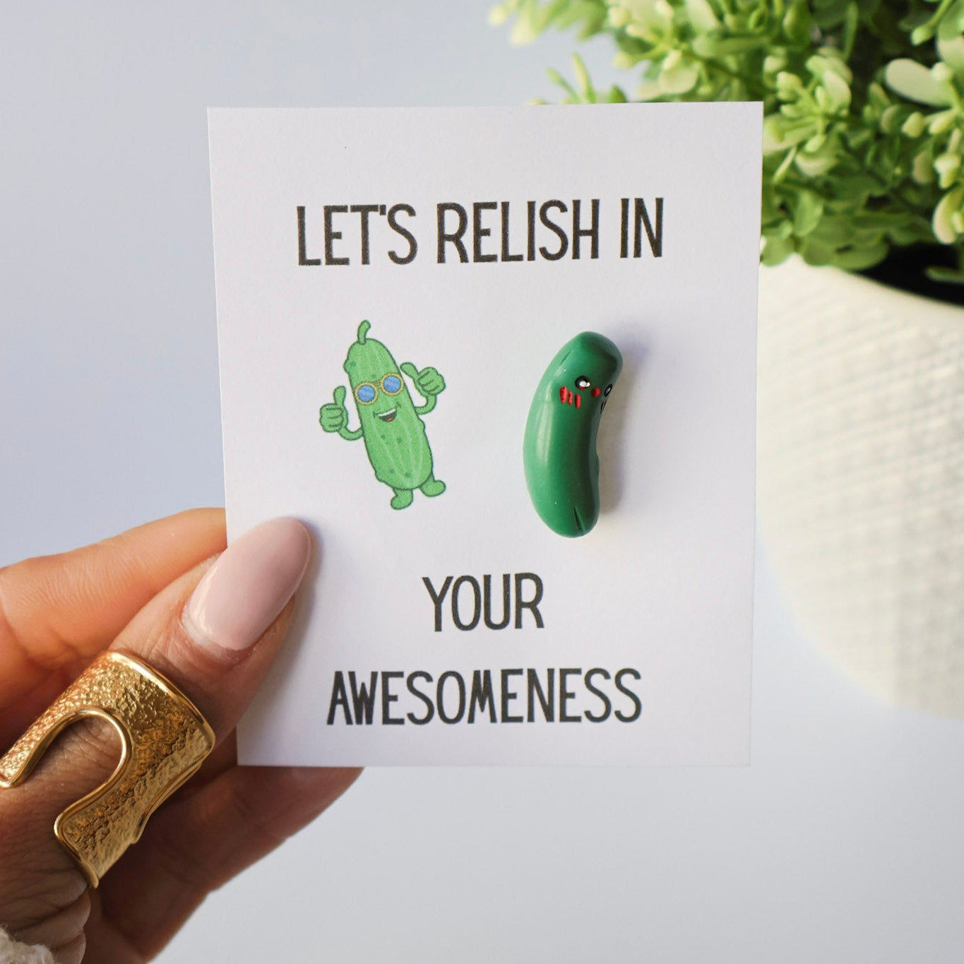 Card with cartoon pickle and charm with text 'Let's relish in your awesomeness' held by a hand.